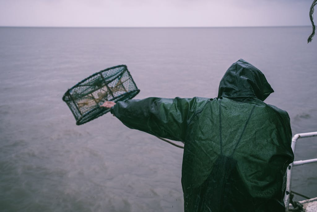 A fisherman in a raincoat throws a crab pot into the sea during a rainy day.