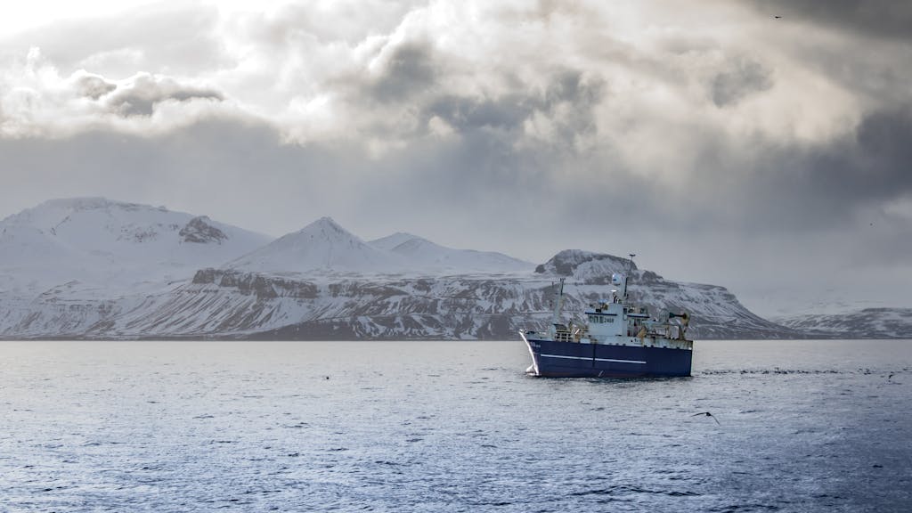 Blue ship navigating icy waters against a backdrop of majestic snow-covered mountains under a dramatic sky.