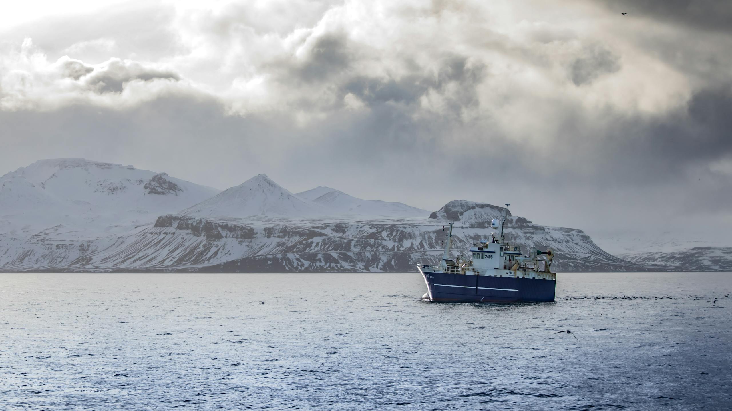 Blue ship navigating icy waters against a backdrop of majestic snow-covered mountains under a dramatic sky.